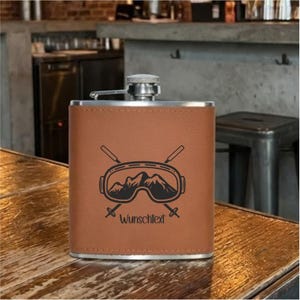 Könnte beinhalten: Eine braune Lederflasche mit silbernem Verschluss. Die Flasche zeigt eine schwarze Grafik von Skibrillen und gekreuzten Skiern mit dem Text "Wunschtext" darunter. Die Flasche ist rechteckig und steht auf einer Holzoberfläche.