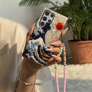 May include: A phone case featuring a wave design in shades of blue and white, with a red sun and floral accents. The case has a multi-camera setup and a pink beaded strap with colorful heart and letter beads.