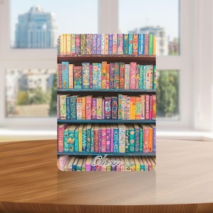 以下が含まれることがあります： カラフルな本棚のデザインが施された、装飾用の長方形のオブジェクト。本棚には、さまざまな色の鮮やかなイラストが描かれた本が並んでいます。「Oliver」という名前が下部に書かれています。背景はぼやけており、窓と都市景観が写っています。