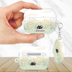 May include: White AirPods case with a floral design and a black cat illustration. The case has the name "Anna" printed on it. A matching keychain is attached. The design features a field of flowers and clouds.