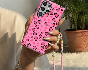 Könnte beinhalten: Eine rosafarbene Smartphone-Hülle mit einem wiederkehrenden Muster aus schwarz umrandeten Herzen. Die Hülle hat eine Aussparung für ein Multikamera-System und ist mit einem Perlenband versehen, das bunte Scheiben und Buchstabenperlen enthält.