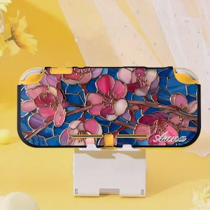 Könnte beinhalten: Eine Switch Lite-Hülle mit einem floralen Design im Glasmalerei-Stil in Rosa und Blau. Die Hülle hat einen schwarzen Rand und gelbe Akzente. Der Name "Anna" ist unten rechts in Kursivschrift geschrieben.