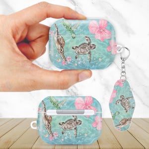Könnte beinhalten: Hellblaues AirPod-Etui und Schlüsselanhänger mit einem Aquarelldesign von Meeresschildkröten, rosa Hibiskusblüten und grünen Meeresschildkrötenflossen. Das Etui ist quadratisch und der Schlüsselanhänger hat eine ovale Form.
