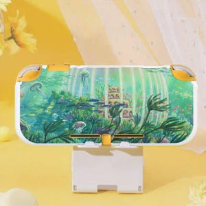 Puede incluir: Una funda blanca y amarilla para Nintendo Switch Lite con una escena submarina que incluye un castillo, peces y medusas. La funda está sostenida por un soporte blanco, con un tema acuático.