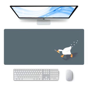 Op de afbeelding: Een grote, rechthoekige bureaumat in een gedempte teal kleur. Een cartoon gans met een mes rent over de mat. Een computertoetsenbord, muis en monitor zijn ook zichtbaar.
