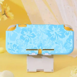 May include: A light blue handheld gaming console case with a floral pattern in a darker shade of blue. The case has yellow accents and is resting on a white stand. The overall design is tropical.