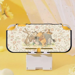 Puede incluir: Una funda amarilla y negra para Nintendo Switch Lite con un estampado floral e ilustraciones de Winnie the Pooh y sus amigos. La funda está sostenida por un soporte blanco.