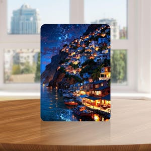 May include: A rectangular art print depicts a coastal village at night. The image shows a hillside dotted with illuminated buildings, a dark blue sea, and boats. The sky transitions from dark blue to a starry night. The print is displayed on a wooden surface.