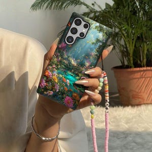 May include: A smartphone case featuring a vibrant floral landscape design with a turquoise river and colorful flowers. The case has a black camera module and a pink beaded strap with the word "LOVE".