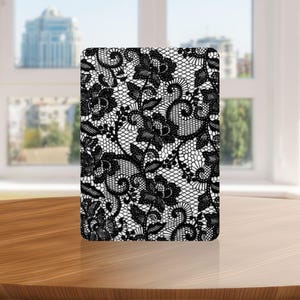 Puede incluir: Una funda rectangular para tableta con un patrón de encaje negro de alto contraste impreso sobre un fondo blanco sólido. El diseño muestra motivos florales intrincados, enredaderas en espiral y una textura de malla fina.