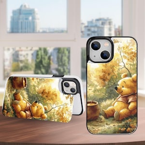 May include: A phone case featuring a watercolor illustration of a yellow bear in a forest setting with a honey pot. The case is white with a black border and a matching design on the front and back.