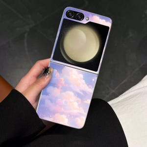 Op de afbeelding: Een opvouwbare smartphonehoes met een pastelkleurig hemelontwerp. De hoes heeft een lichtpaarse basis met roze en witte wolken en sterren. Het scherm toont een grote, heldere, gele bol tegen een zwarte achtergrond. De telefoon wordt in een hand gehouden.