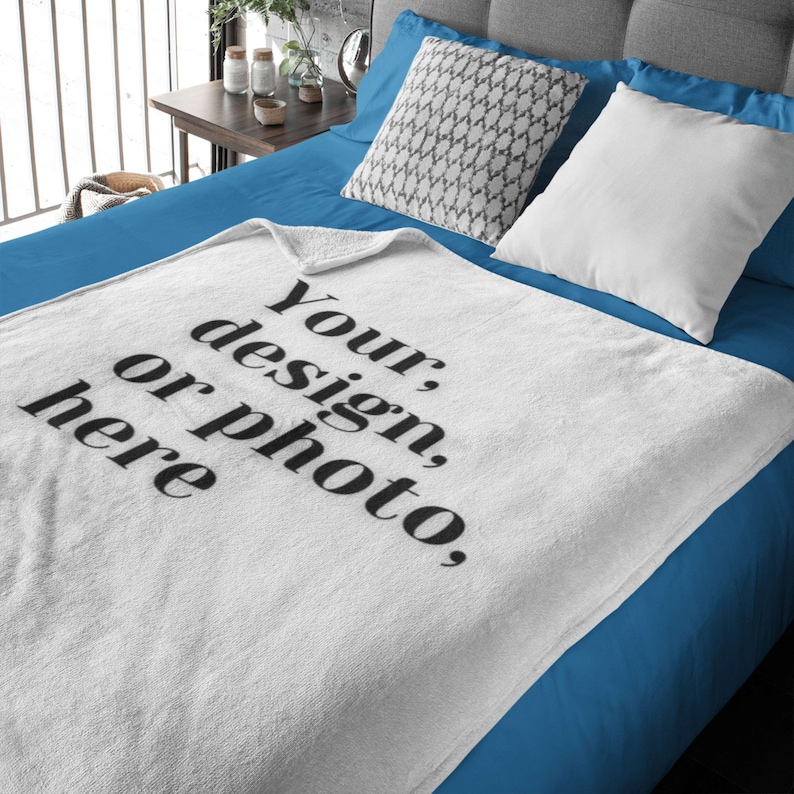 May include: White blanket with the text "Your, design, or photo, here" in black. The blanket is folded on a blue bed with white and patterned pillows. The blanket is made of a soft material.