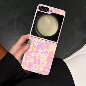 Puede incluir: Una funda plegable para smartphone con un estampado floral en rosa, morado y crema. La funda abierta revela una pantalla que muestra una esfera clara. El teléfono tiene una cámara y una pequeña abertura circular en la parte superior.