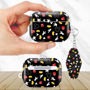 May include: Black AirPods case and keychain with a repeating pattern of Mickey Mouse gloves, ears, and shorts in white, red, and yellow. The case is held in a hand, and another case and keychain are on a wooden surface.