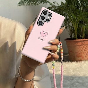 Puede incluir: Una funda de teléfono rosa claro con un corazón y la palabra "Smile" impresa. La funda tiene un sistema multicámara y una correa de cuentas. La correa incluye cuentas en forma de corazón y la palabra "Love".