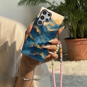 May include: A smartphone case with a landscape design in shades of blue and gold. The case has a strap with colorful beads and the word "LOVE". The phone has multiple camera lenses.