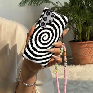 May include: A black and white phone case featuring a bold, spiraling hypnotic pattern. The case has cutouts for a multi-lens camera system. Attached to the side is a decorative beaded phone strap with colorful, small, flat, circular beads.