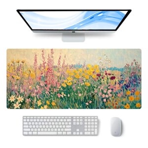 Op de afbeelding: Een rechthoekige bureaumat met een levendig bloemenschilderij in tinten geel, roze en paars. Een wit toetsenbord, muis en computermonitor zijn boven en onder de mat geplaatst. Het kunstwerk toont een veld met wilde bloemen.
