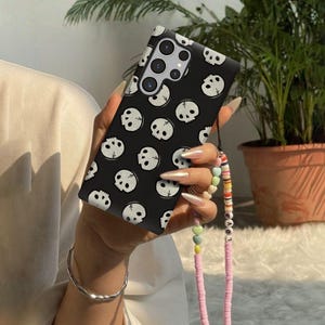 May include: Black phone case with a repeating pattern of white skull illustrations. The case has a strap with colorful beads and the word "LOVE". The phone case is held in a person's hand.
