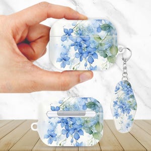 Op de afbeelding: Witte AirPod-hoes en sleutelhanger met een blauw bloemmotief en groene bladeren. De hoes wordt door een hand vastgehouden en toont de bloemen in aquarel. De sleutelhanger heeft een vergelijkbaar ontwerp.