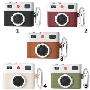May include: Five different colored camera-shaped phone cases with a silver carabiner. The cases are black, red, brown, white, and green. Each case has a white front with a black lens and the text "Designed in California" printed on the front.