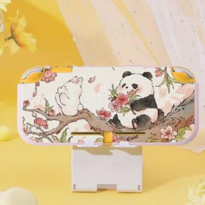 May include: A pink and yellow Nintendo Switch Lite case featuring a whimsical illustration of a panda and a rabbit on a tree branch with pink flowers. The case is resting on a white charging stand.