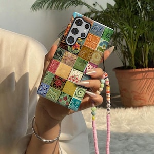 Puede incluir: Una funda de teléfono colorida con un diseño de mosaico. La funda presenta una variedad de baldosas cuadradas en tonos verdes, amarillos, rosas y azules, con patrones florales y geométricos. Una correa de cuentas rosas con la palabra "LOVE" está adjunta.