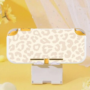 May include: A white and yellow handheld gaming console case with a beige leopard print design. The case is resting on a white stand, with a yellow background.