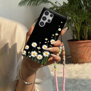 May include: Black phone case with a daisy floral design. The case features white daisies with yellow centers and green stems against a black background. A pink and beaded lanyard is attached to the phone case.