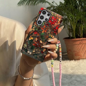 Op de afbeelding: Een kleurrijke telefoonhoes met een bloemig glas-in-lood ontwerp. De hoes heeft rode, oranje, groene en bruine tinten. Een roze kralenriem met kleurrijke kralen en het woord "LOVE" is bevestigd.