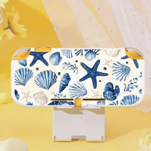 May include: A white and yellow Nintendo Switch Lite case featuring a pattern of blue seashells, starfish, and aquatic plants. The case has yellow accents on the corners and buttons. The design is set against a cream-colored background.