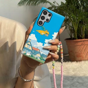 May include: A blue phone case featuring a cartoon dragon flying in a blue sky with clouds. The case also depicts houses and a strap with colorful beads and the word "LOVE".