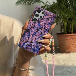 Op de afbeelding: Een telefoonhoesje met een bloemenpatroon en een bandje. De hoes heeft een patroon van kleine paarse en blauwe bloemen. Het bandje is roze met kleurrijke kralen en het woord "LOVE".