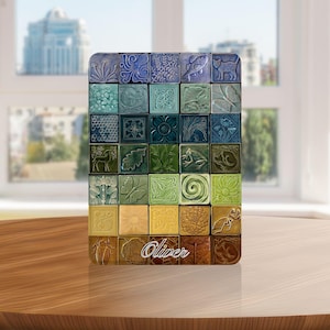 Puede incluir: Una colección de baldosas cerámicas cuadradas en varios tonos de azul, verde y amarillo, cada una con un diseño en relieve único de plantas, animales y patrones geométricos. El nombre "Oliver" está escrito en las baldosas inferiores.