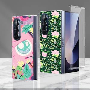 May include: Samsung Galaxy Z Fold phone cases. One case features a pink background with a cartoon eye and floral elements. The other case has a green background with a floral pattern. A clear case is also shown.