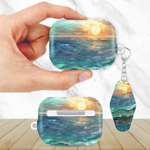 May include: AirPods case and keychain featuring a sunset over the ocean design. The case is rectangular with rounded edges, and the keychain is an oval shape. The design includes shades of blue, green, yellow, and orange.