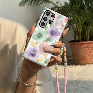 May include: A floral phone case with a pastel watercolor design. The case features purple, pink, and green flowers with gold accents on a white background. A pink beaded strap with the word "LOVE" is attached.
