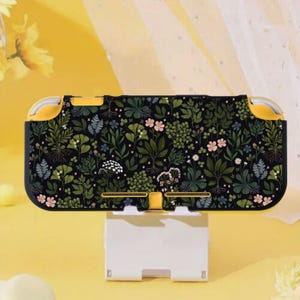 Könnte beinhalten: Eine schwarze und gelbe Nintendo Switch Lite-Hülle mit einem botanischen Muster aus grünen Blättern und rosa Blüten. Die Hülle steht auf einem weißen Ständer vor einem gelben Hintergrund. Die Hülle hat gelbe Akzente.