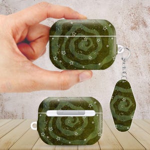 May include: A dark olive green protective case for wireless earbuds featuring a swirling wave pattern with small white stars and speckles. The set includes a matching oval-shaped keychain fob attached by a silver metal ring and chain.