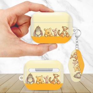 May include: Yellow and orange AirPods case featuring Winnie the Pooh characters: Eeyore, Winnie the Pooh, Piglet, and Tigger. Includes a matching keychain with the same characters. The case is held by a hand.