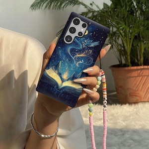 May include: A dark blue phone case with a fantasy book design. The case features an open book with glowing pages, a celestial background, and a strap with colorful beads. The phone case is held in a person's hand.