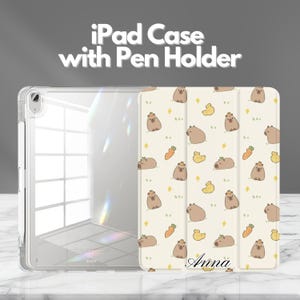 Peut inclure: Coque transparente pour iPad avec porte-stylo et housse à motifs. La housse présente un fond crème avec des capybaras, des carottes et des canards jaunes de dessins animés. Le texte "iPad Case with Pen Holder" est en haut. Le nom "Anna" est en bas.