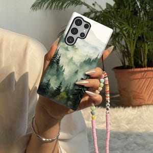 May include: A white smartphone case featuring a misty forest scene with green and gray tones. The case has a black strap with colorful beads and the word "LOVE".