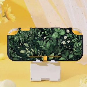 Può includere: Una custodia per Nintendo Switch Lite con un design botanico verde e bianco, incorniciata in giallo e nero. Poggia su un supporto di ricarica bianco. Il design presenta foglie e piccoli fiori bianchi.