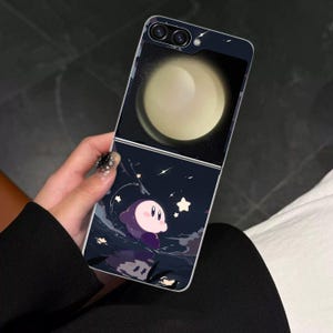 Puede incluir: Funda de teléfono con un diseño en dos partes. La sección superior muestra un orbe amarillo brillante sobre un fondo oscuro. La sección inferior presenta un personaje de dibujos animados en morado y rosa, sobre un cielo estrellado.