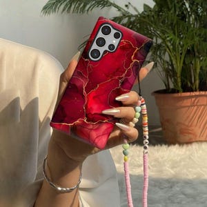 May include: A smartphone case featuring a deep red and burgundy marble pattern with thin gold veins. The case has a cutout for a multi-lens camera system and is attached to a beaded phone strap with colorful discs and the word LOVE spelled out.