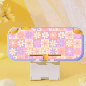 May include: A lavender and yellow Nintendo Switch Lite case featuring a daisy floral pattern. The case has a checkerboard design with pink, purple, and peach-coloured flowers. The case is resting on a white stand.