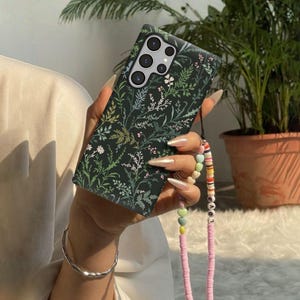May include: A dark green smartphone case featuring a botanical pattern of white, light green, and pink wildflowers and ferns. The case includes a multi-colored beaded phone strap with pastel-colored beads and letter beads spelling out LOVE.