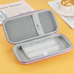 以下が含まれることがあります： 開いたラベンダー色のジッパー付きキャリングケース。グレーのメッシュインテリアと白い長方形の収納コンパートメントがあります。このケースは、電子機器やアクセサリーを収納するように設計されています。ケースは黄色の表面にあります。